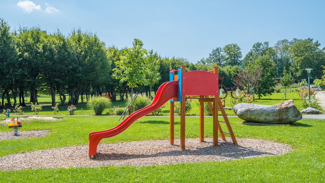Ein Spielplatz mit einer roten Rutsche steht auf einer grünen Wiese. Im Hintergrund sind Bäume und ein klarer Himmel zu sehen. | © Helmut Schweighofer