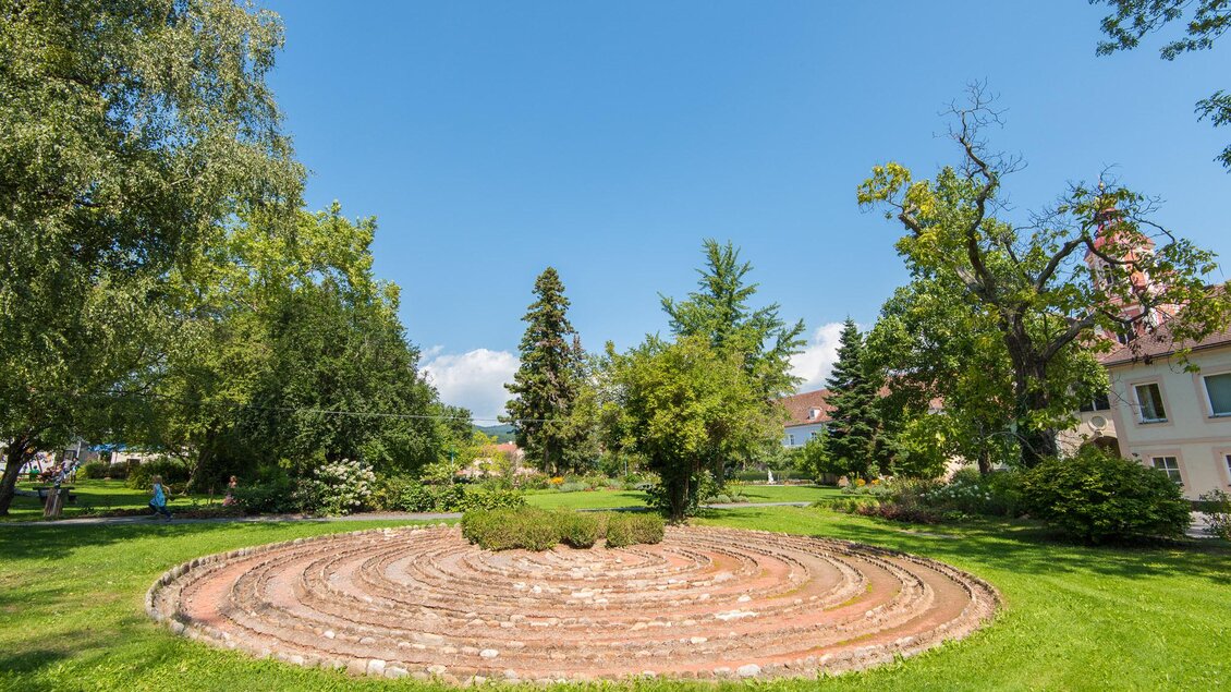 Ein runder Garten mit einer spiralförmigen Steinbepflanzung. Üppiges Grün und Bäume umgeben die Fläche unter einem klaren blauen Himmel. | © Helmut Schweighofer