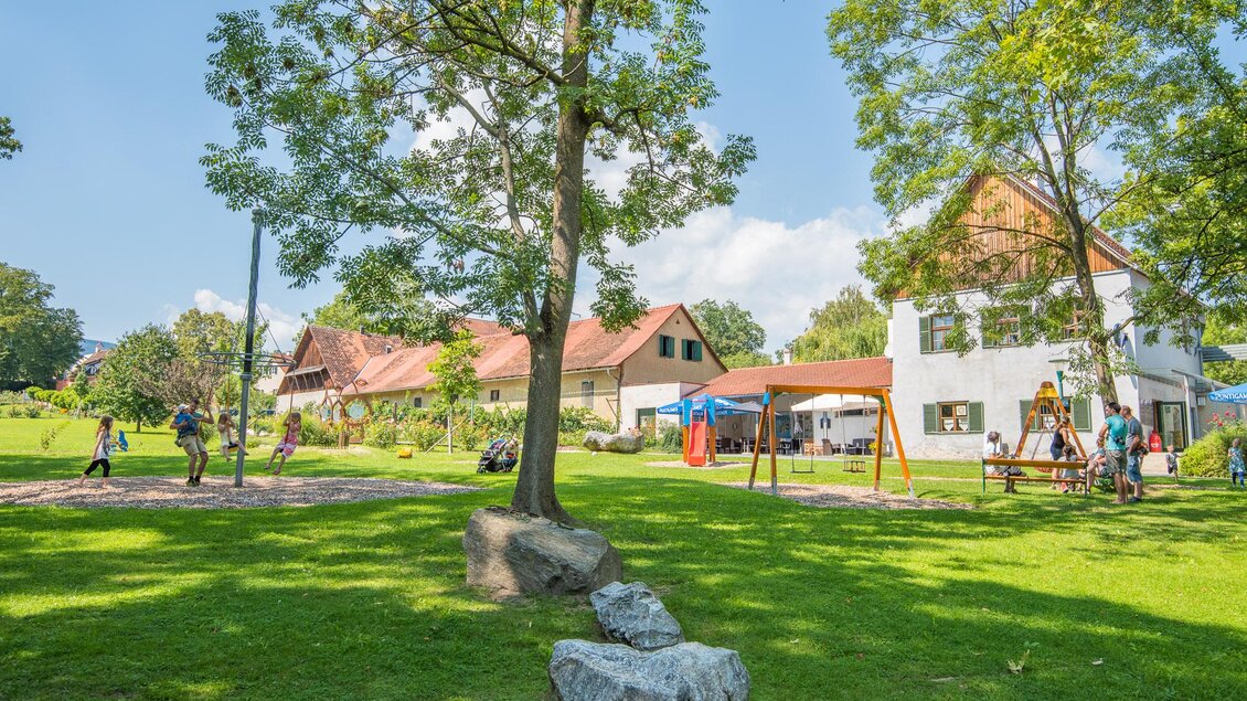 Ein schöner Garten mit viel Grün und einem großen Baum. Im Hintergrund sind ein Spielplatz und einige Gebäude zu sehen. | © Helmut Schweighofer