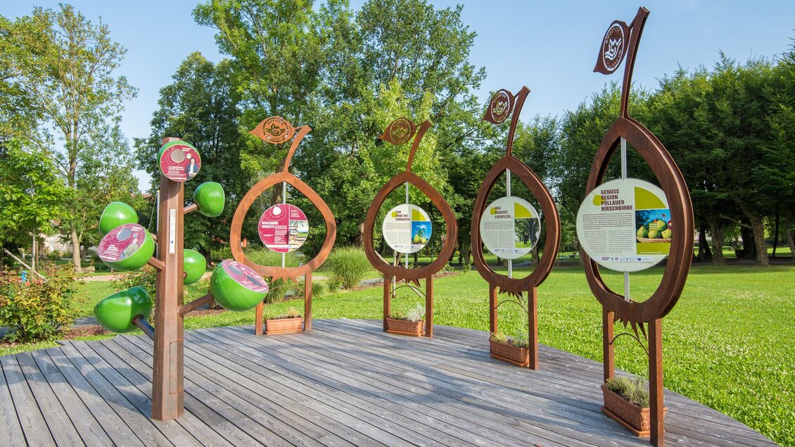 Eine Holzterrasse mit mehreren informativen Schildern in organischen Formen. Im Hintergrund sind grüne Bäume und ein klarer Himmel zu sehen. | © Helmut Schweighofer