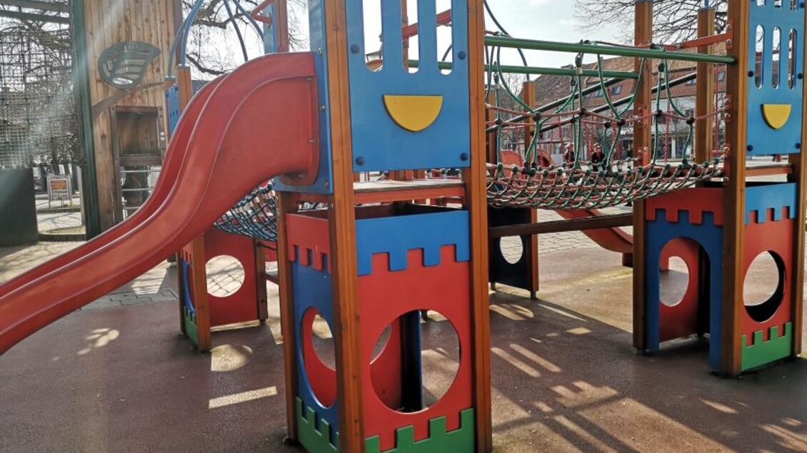 Ein bunter Spielplatz mit einer roten Rutsche und einem Klettergerüst. Im Hintergrund sind Holzstrukturen und Bäume zu sehen. | © Petra Stossier