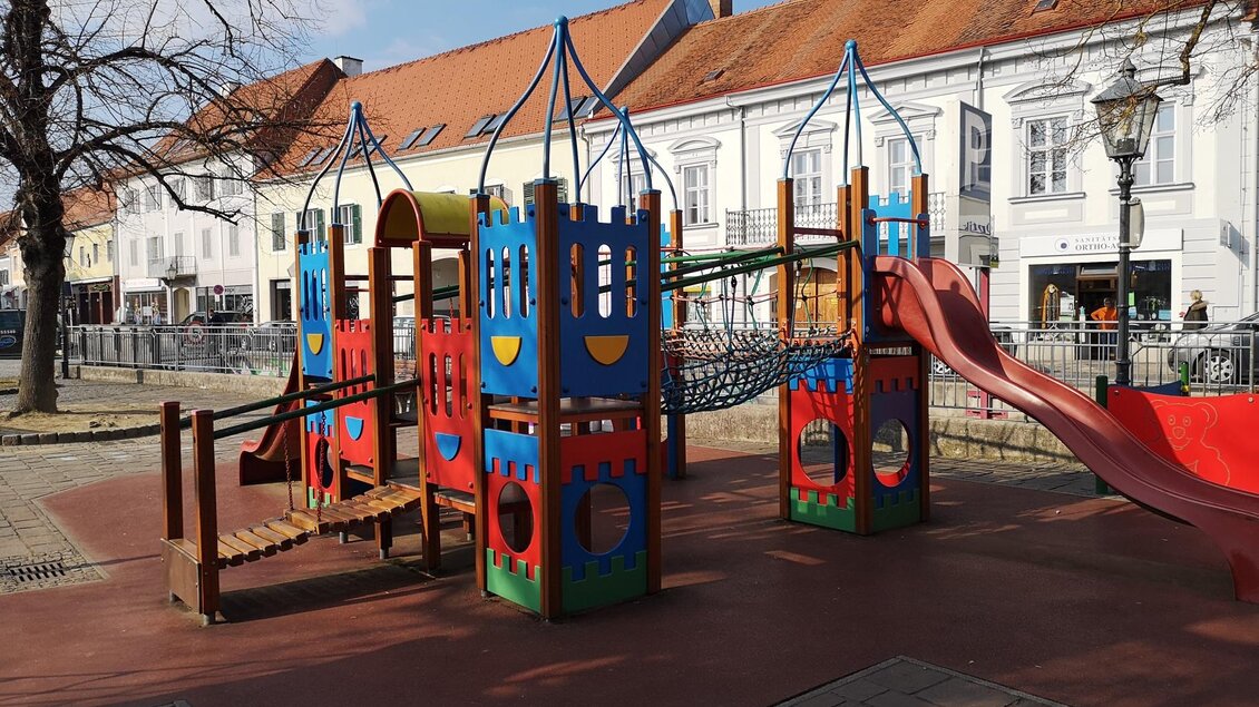 Ein bunter Spielplatz mit Spielgeräten und einer Rutsche. Im Hintergrund sind helle Gebäude und Bäume zu sehen. | © Petra Stossier