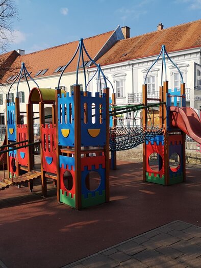 Ein bunter Spielplatz mit Spielgeräten und einer Rutsche. Im Hintergrund sind helle Gebäude und Bäume zu sehen. | © Petra Stossier