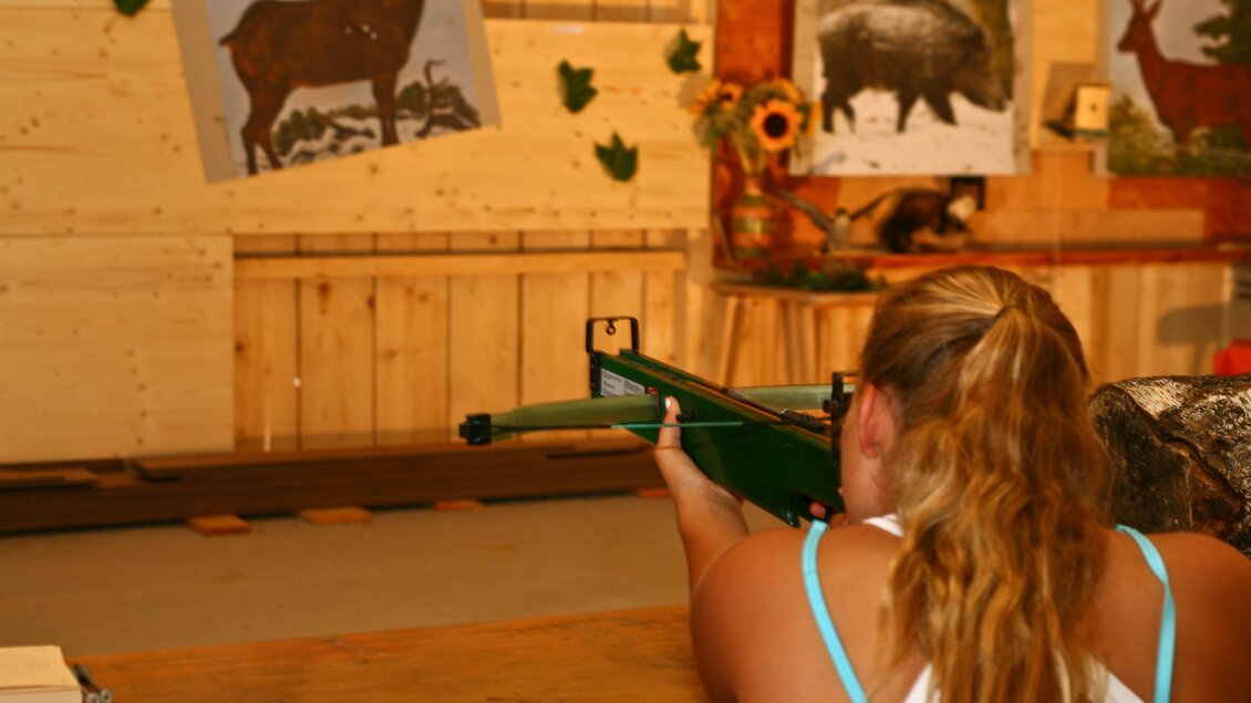 Ein Mädchen zielt mit einem Gewehr auf Zielbilder von Tieren. Der Hintergrund zeigt eine rustikale Umgebung mit Holzpanelen. | © Bärenwirt