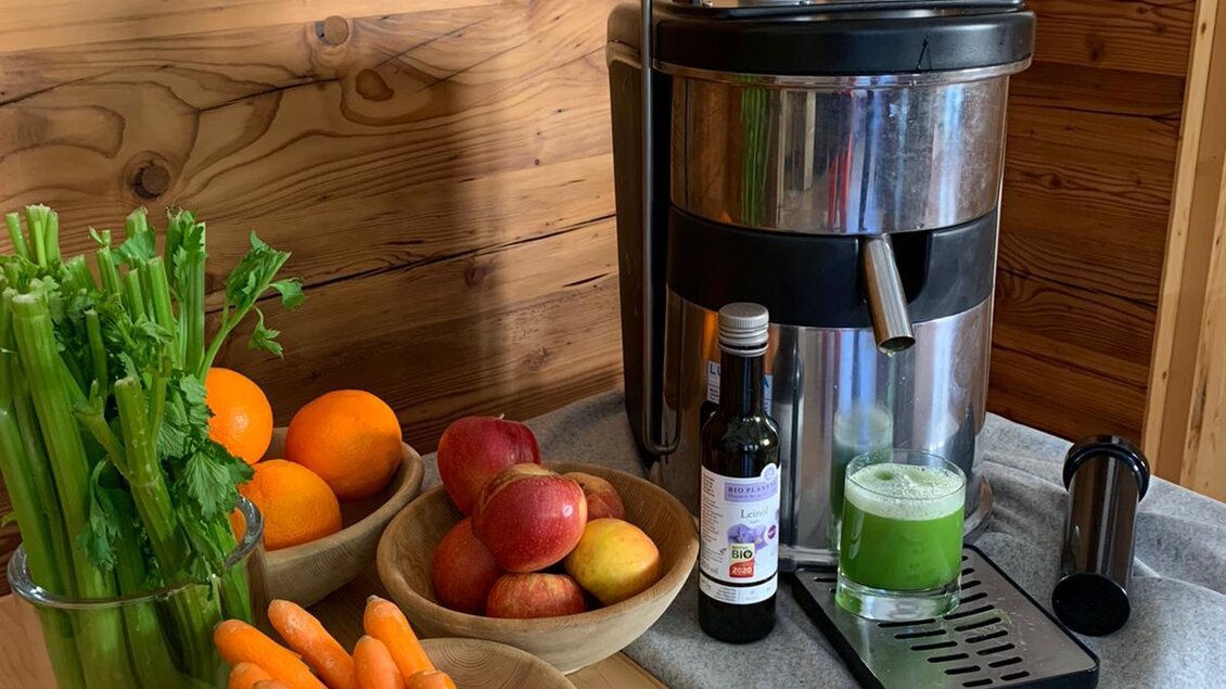 Eine Saftpresse steht in einer Küche neben frischem Obst und Gemüse. Auf dem Tisch liegen Karotten, Sellerie, Äpfel und Orangen, sowie ein Glas grüner Saft. | © baerenwirt.com