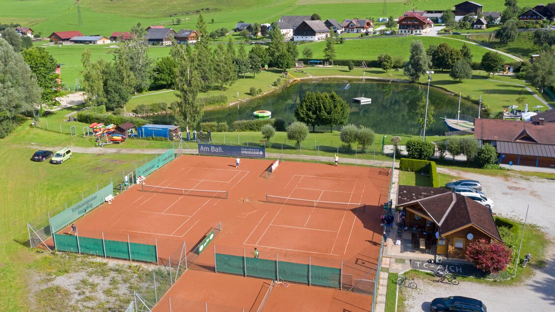Eine Tennisanlage mit roten Sandplätzen und einem kleinen Teich in der Nähe. Umgeben von einer grünen Landschaft und Wohngebäuden. | © Raffalt Herbert
