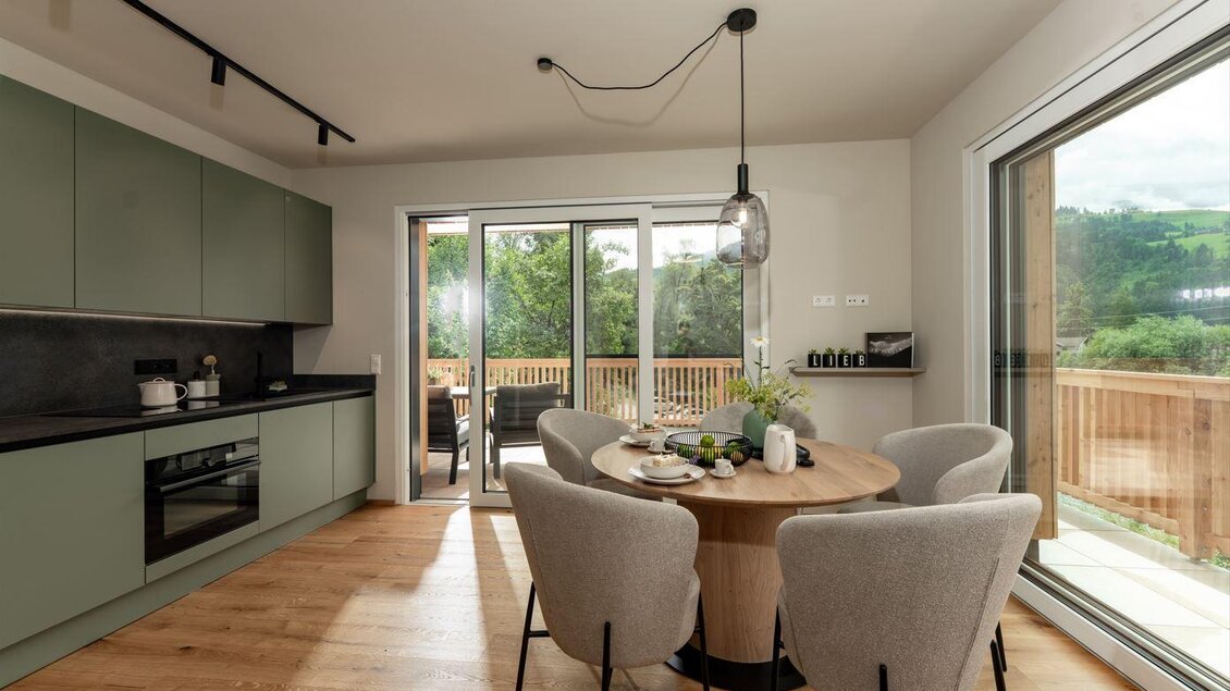 Offene, moderne Küche in sanftem Grün mit Essbereich und großem Rundtisch, davor große Glasfront mit Zugang auf die Terrasse und Blick ins Grüne. | © Bärenwirt