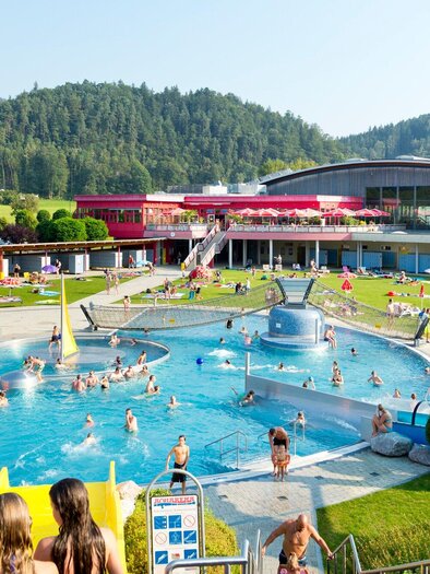 Ein großer Schwimmbereich mit vielen Menschen und einem Rutschenbereich im Freizeitpark Frohnleiten. Im Hintergrund sind grüne Hügel und ein modernes Gebäude zu sehen. | © TV Region Graz - Rene Vidalli