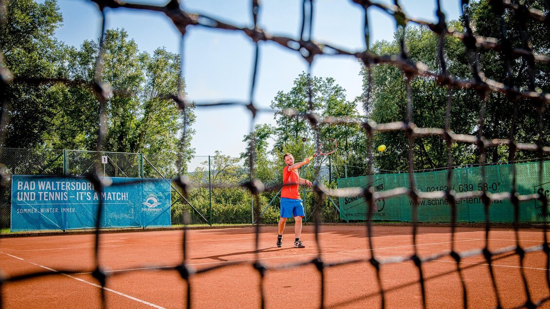 Ein Mann spielt Tennis auf einem roten Tennisplatz. Im Hintergrund sind Bäume und eine Werbetafel sichtbar. | © TVB Bad Waltersdorf