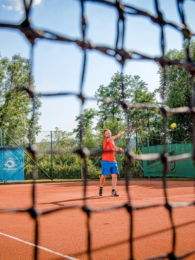 Ein Mann spielt Tennis auf einem roten Tennisplatz. Im Hintergrund sind Bäume und eine Werbetafel sichtbar. | © TVB Bad Waltersdorf