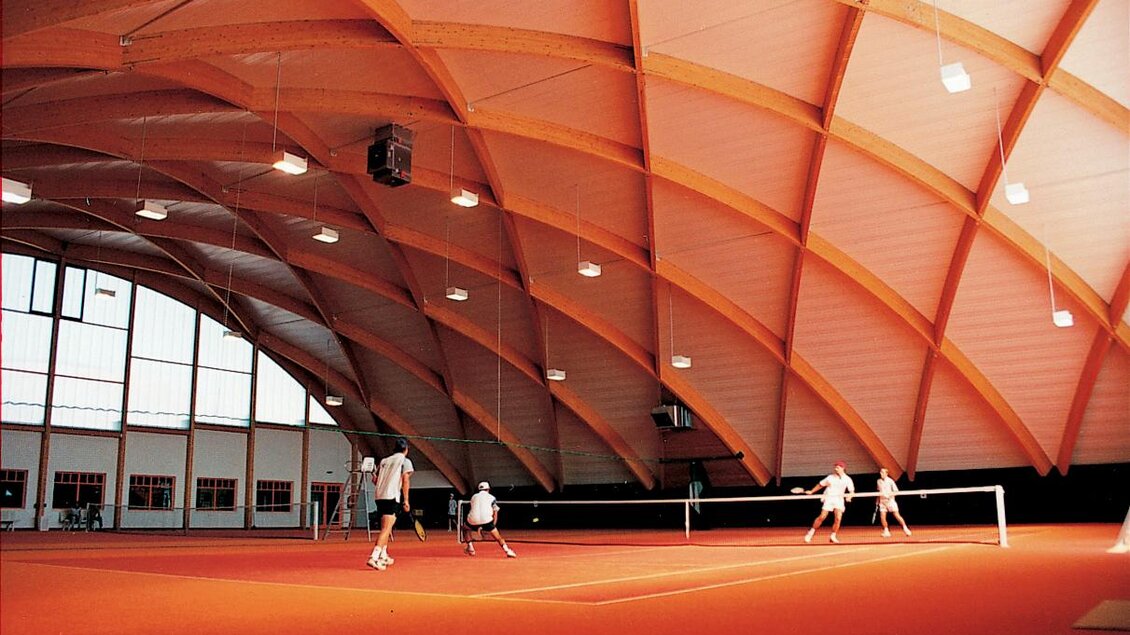 Eine Tennishalle mit einem großen, gewölbten Dach und orangefarbenem Boden. Auf dem Platz spielen mehrere Personen Tennis. | © Mias Photoart