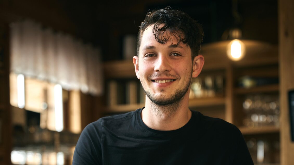 Ein junger Mann lächelt freundlich hinter der Bar. Im Hintergrund sind Gläser und eine gemütliche Atmosphäre zu sehen. | © Nationalpark Gesäuse