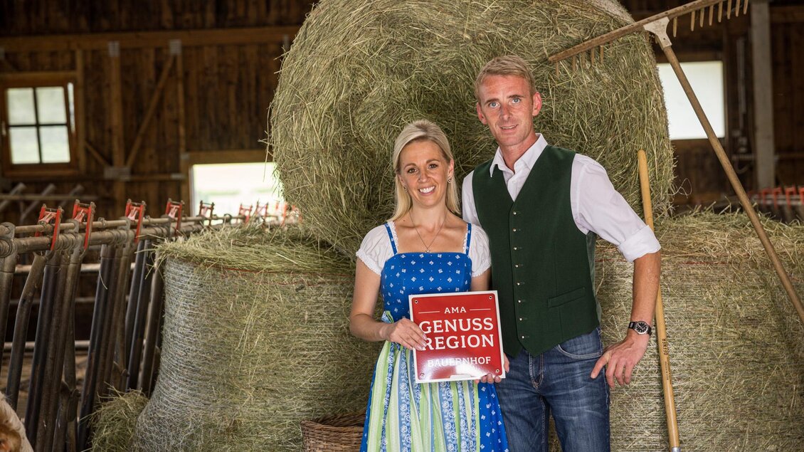 Ein Paar in traditioneller Kleidung steht in einem Bauernhof mit Heuballen im Hintergrund. Sie halten ein Schild mit der Aufschrift „Genussregion“. | © Netzwerk Kulinarik wildbild