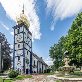 St. Barbara Kirche in Bärnbach mit gestaltetem Kirchturm, Brunnen und Bäumen unter blauem Himmel | © Die Abbilderei