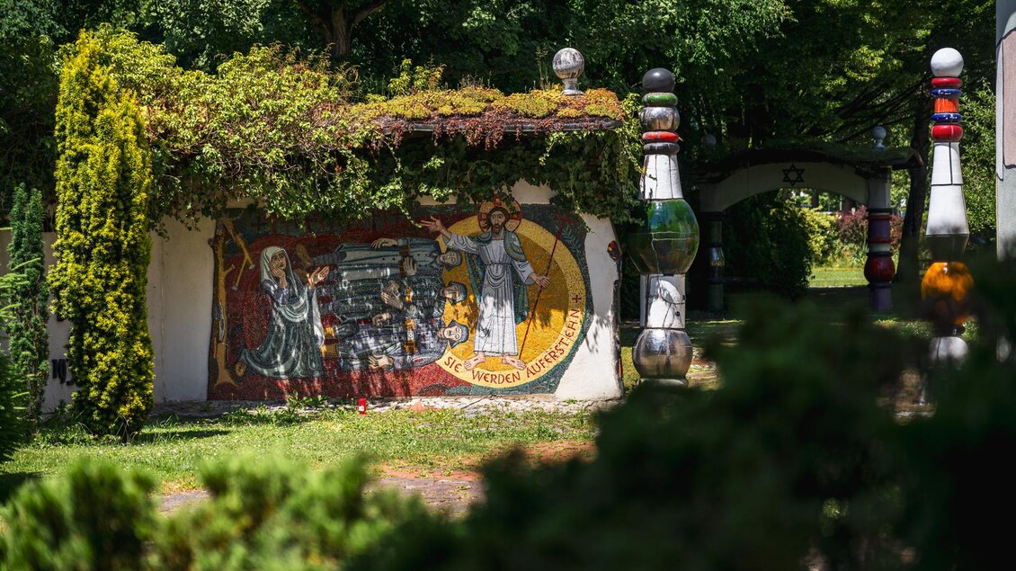 Bemalte Wand und farbige Säulen im begrünten Außenbereich der von Hundertwasser gestalteten St. Barbara Kirche | © Region Graz_Bernd Grosseck