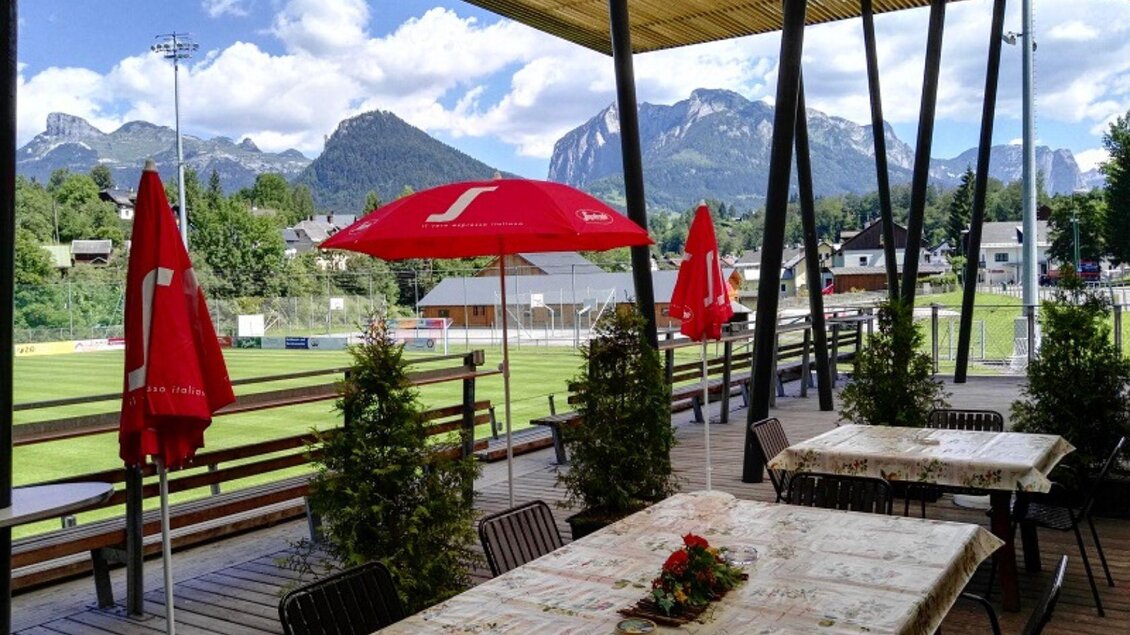Eine Terrasse mit Tischen und Sonnenschirmen bietet einen Blick auf ein Fußballfeld und beeindruckende Berge im Hintergrund. Die Umgebung ist grün und einladend. | © Stadionstüberl Bad Aussee (c) Jochen Kreidl