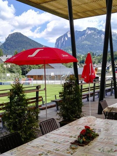 A terrace with tables and umbrellas offers a view of a soccer field and impressive mountains in the background. The surroundings are green and inviting. | © Stadionstüberl Bad Aussee (c) Jochen Kreidl