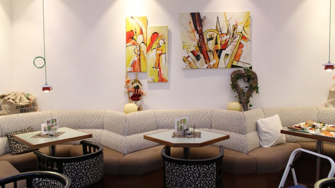 Heller Gastraum des Stadtcafés Voitsberg mit Eckbank, Tischen, Stühlen und farbenfrohen Wandbildern. | © Stadtcafe - Uhl