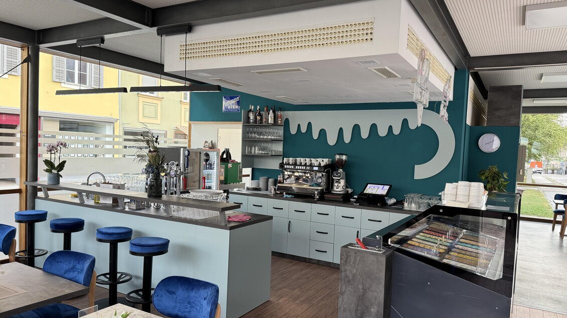 Ein modernes Café mit einer eleganten Bar und blauen Stühlen. Die Wände sind in sanften Farben dekoriert, und es gibt gewellte Designelemente. | © Mesut Tunc