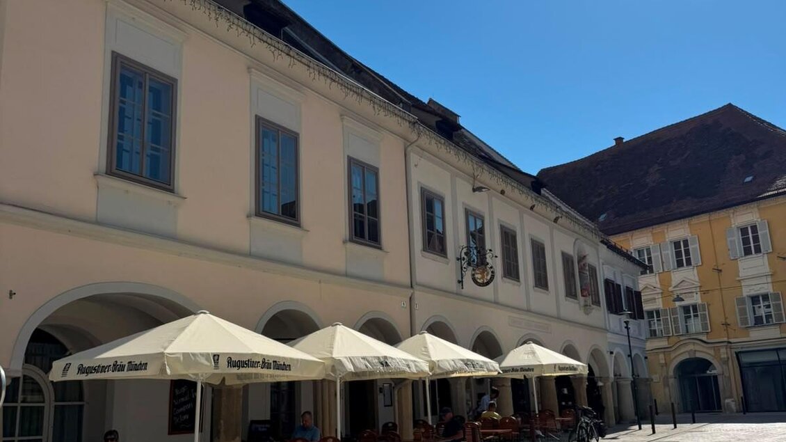 Ein sonniger Platz mit einem Restaurant und Terrassen unter Sonnenschirmen. Die Gebäude im Hintergrund zeigen eine charmante Architektur. | © Thermen-&Vulkanland JT