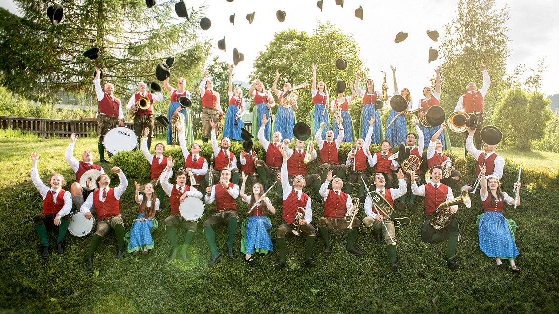 Eine fröhliche Gruppe von Musikern steht im Freien und jubelt. Sie tragen bunte Trachten und werfen ihre Hüte in die Luft. | © Shooting Star