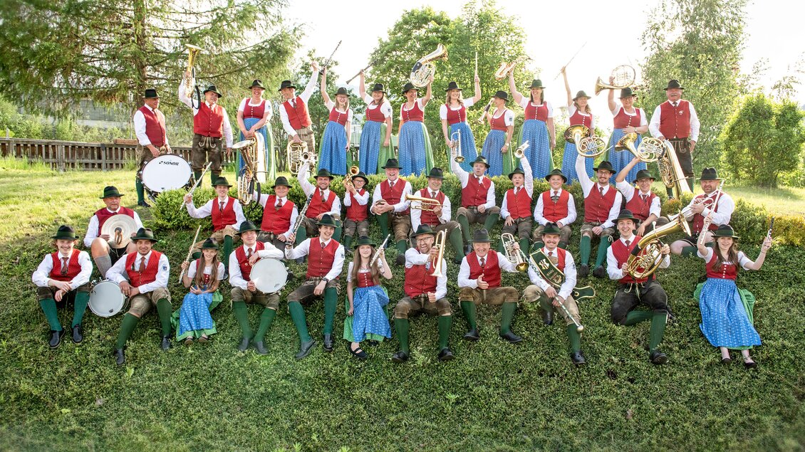 Eine großen Musikgruppe sitzt und steht im Freien. Die Mitglieder tragen traditionelle Trachten in Rot und Blau. | © Shooting Star