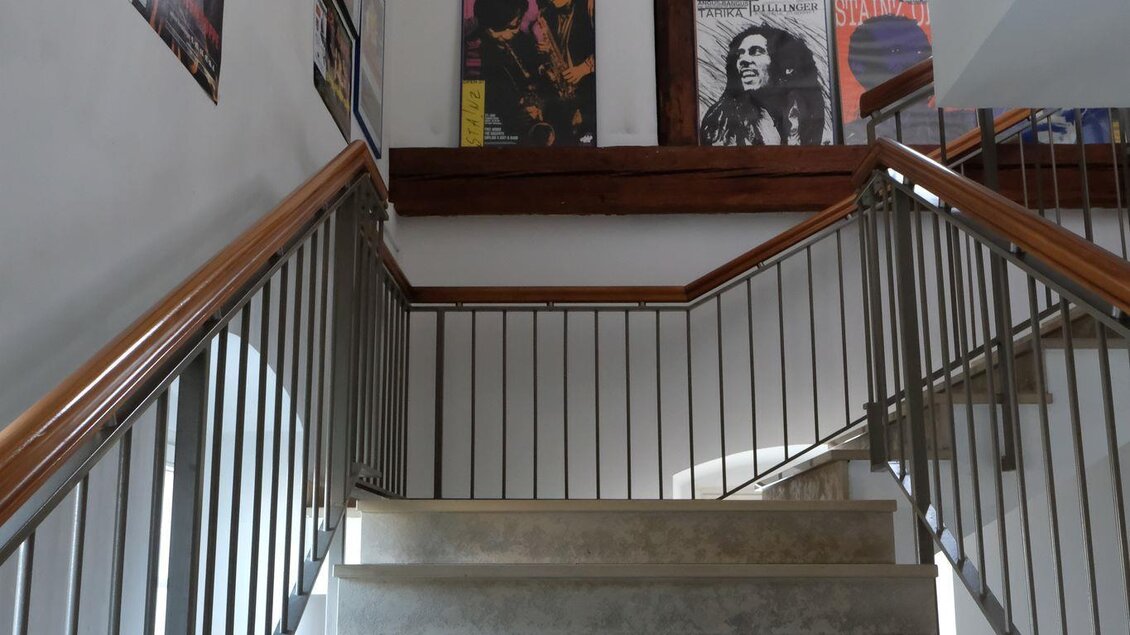 Eine Treppe mit hellen Stufen und einem Handlauf aus Holz führt nach oben. An der Wand hängen verschiedene Bilder und Poster. | © Helmut Fladenhofer