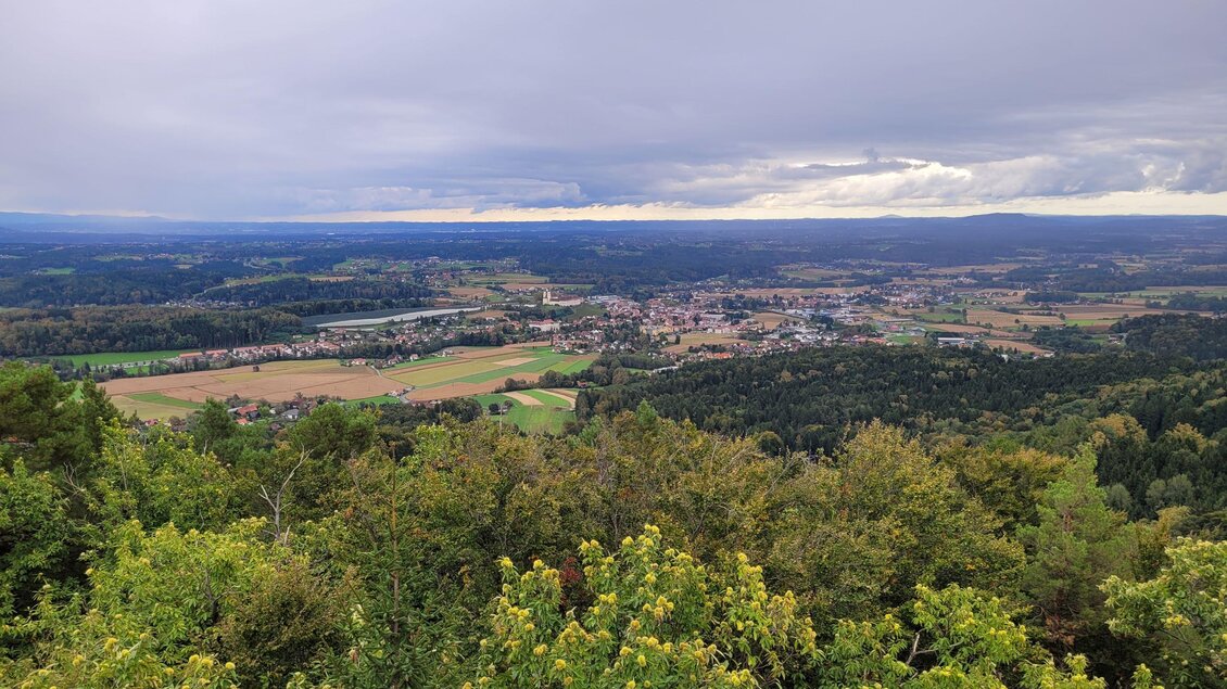 Eine weitläufige Landschaft mit grünen Bäumen und Feldern. Im Hintergrund ist eine kleine Stadt unter einem bewölkten Himmel zu sehen. | © TV Südsteiermark - Irene Löschnig