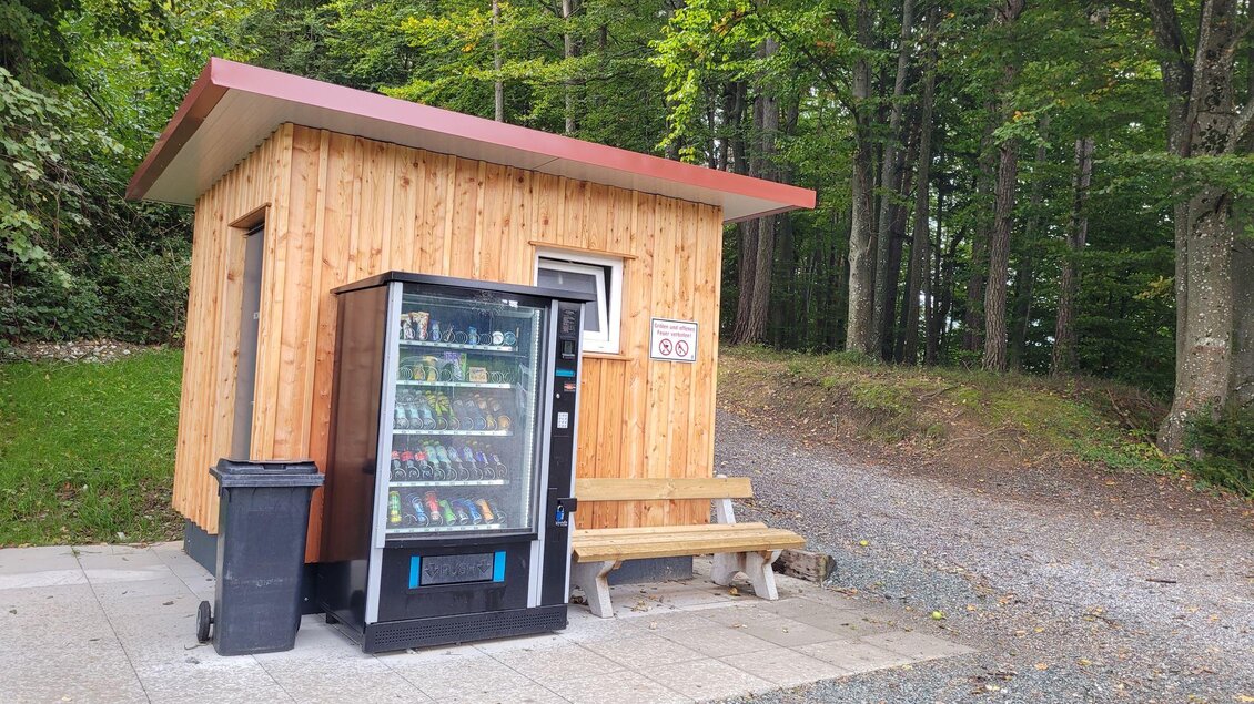 Ein kleines Holzgebäude steht im Wald neben einem Verkaufsautomaten. Draußen gibt es eine Bank zum Sitzen. | © TV Südsteiermark - Irene Löschnig