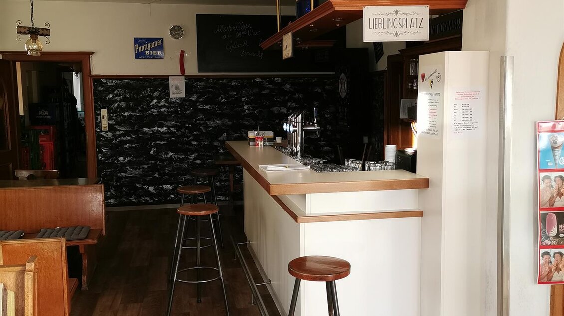 Eine gemütliche Bar mit Holzfußboden und einem hellen Thekenbereich. Im Hintergrund sind Stühle und dekorative Elemente zu sehen. | © Stambach-Wirt