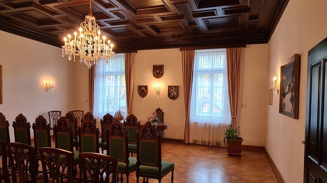 Ein eleganter Raum mit einem dekorativen Holzboden und einer klassischen Decke. Im Hintergrund sind ein Kronleuchter und Stühle in grüner Polsterung zu sehen. | © Stephanie Bor