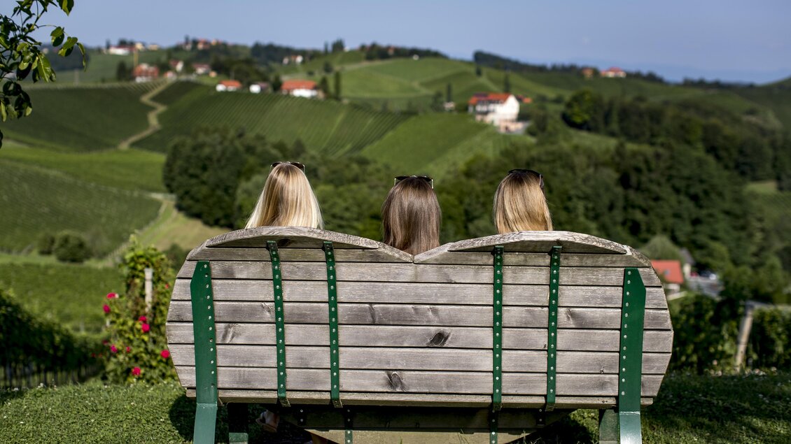 Drei Frauen sitzen auf einer Holzbank und blicken auf eine grüne Weinlandschaft. Im Hintergrund sind Hügel und einige Häuser sichtbar. | © TV Südsteiermark | Tom Lamm