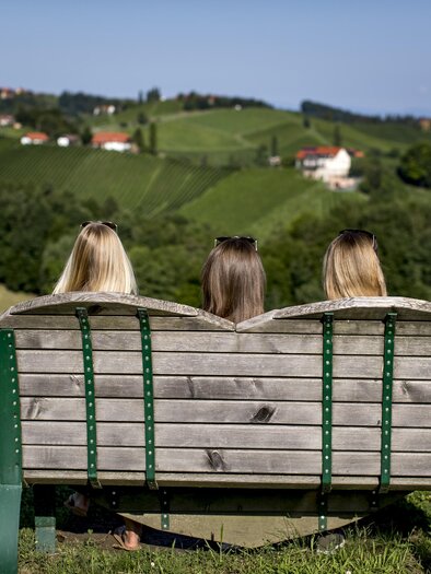 Drei Frauen sitzen auf einer Holzbank und blicken auf eine grüne Weinlandschaft. Im Hintergrund sind Hügel und einige Häuser sichtbar. | © TV Südsteiermark | Tom Lamm