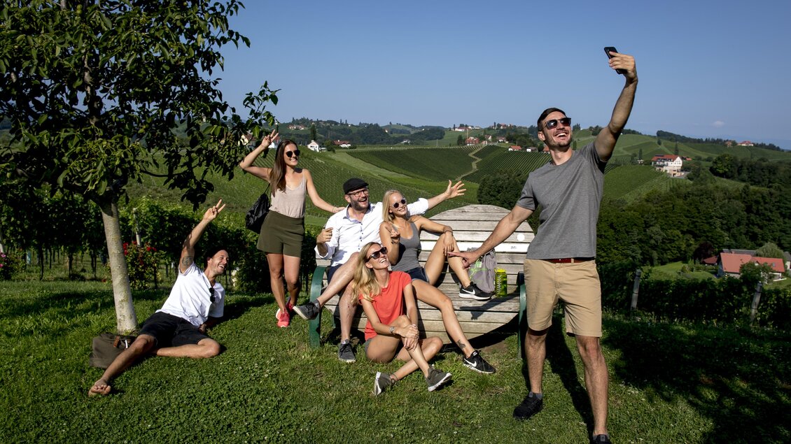 Eine Gruppe von Freunden posiert fröhlich für ein Selfie auf einer grünen Wiese. Im Hintergrund sind sanfte Hügel und ein klarer blauer Himmel zu sehen. | © TV Südsteiermark | Tom Lamm