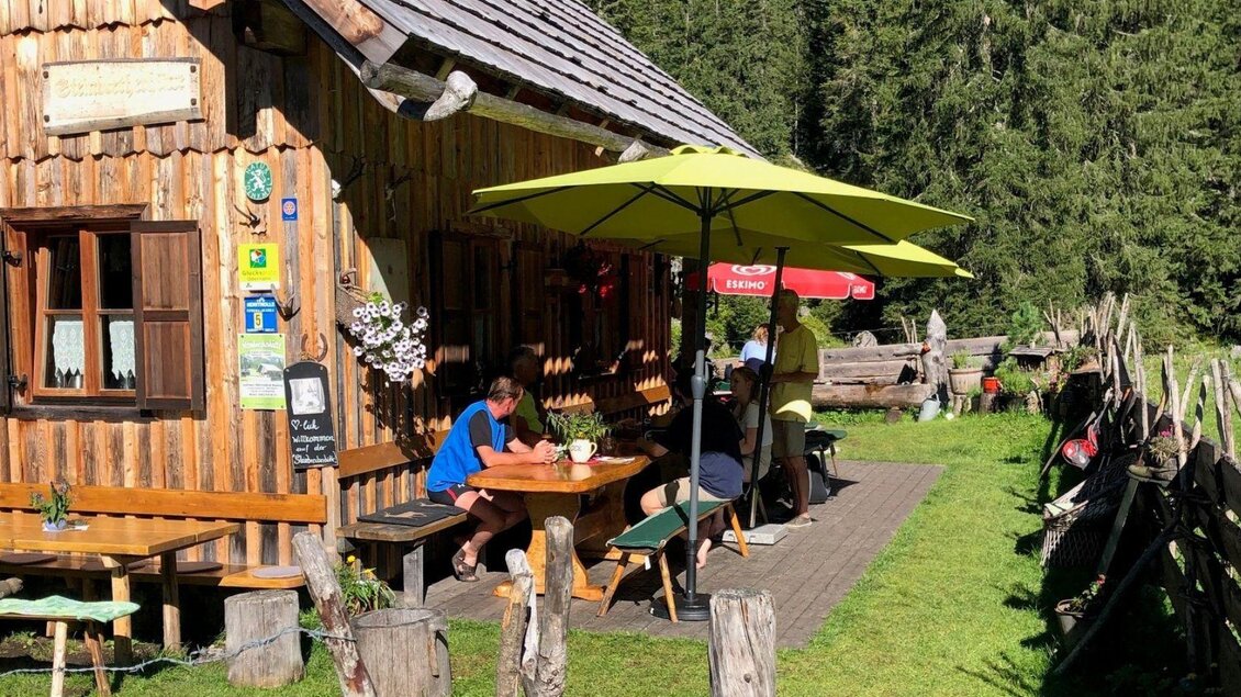 Die charmante Steinbrecherhütte liegt eingebettet von kargen Gebirgszügen auf der grünen Hochebene der Ödernalm in Bad Mitterndorf. Vor der Hütte sitzen ein paar Wanderer und genießen kühle Getränke sowie eine Jause in der idyllischen Umgebung. | © Sabine Carstensen