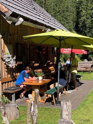 Die charmante Steinbrecherhütte liegt eingebettet von kargen Gebirgszügen auf der grünen Hochebene der Ödernalm in Bad Mitterndorf. Vor der Hütte sitzen ein paar Wanderer und genießen kühle Getränke sowie eine Jause in der idyllischen Umgebung. | © Sabine Carstensen