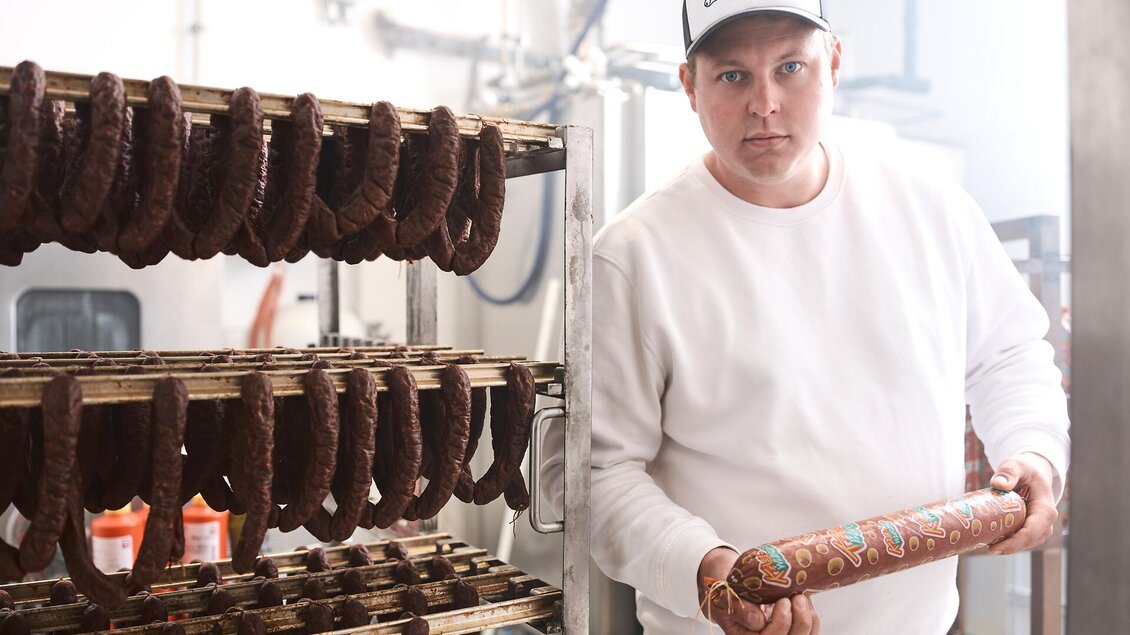 Ein Mann hält eine Rolle Wurst und steht neben einem Regal mit getrockneter Wurst. Die Umgebung wirkt wie eine Lebensmittelproduktionsanlage. | © Nationalpark Gesäuse