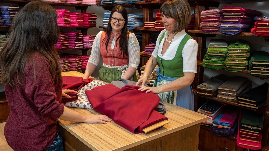 Drei Frauen stehen an einem Tisch in einem Stoffladen. Sie diskutieren und betrachten verschiedene Stoffe, die ordentlich auf Regalen im Hintergrund angeordnet sind. | © Landmarkt, Christian Wöhr
