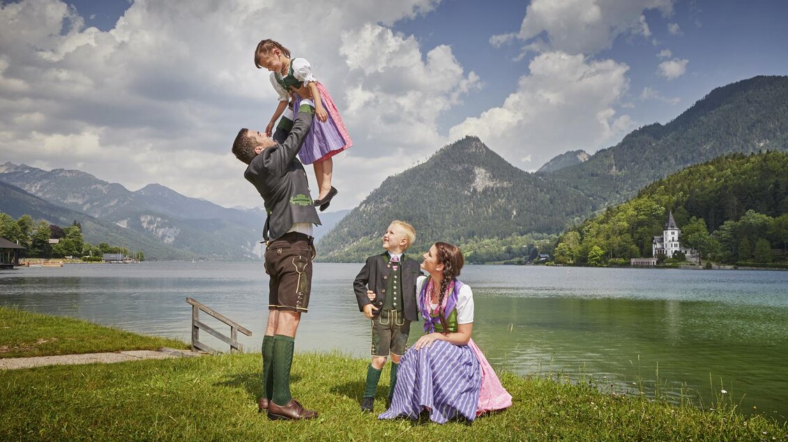 Eine Familie in traditioneller Tracht steht am Ufer eines Sees mit Bergen im Hintergrund. Ein Erwachsener hebt ein Kind in die Luft, während zwei weitere Kinder zuschauen. | © photoInStyle, Birgit Steinberger