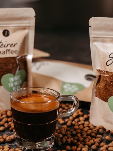A glass of fresh coffee stands in front of two packages of Steirer Kaffee. Coffee beans are scattered across the table surface. | © Region Graz - Mias Photoart
