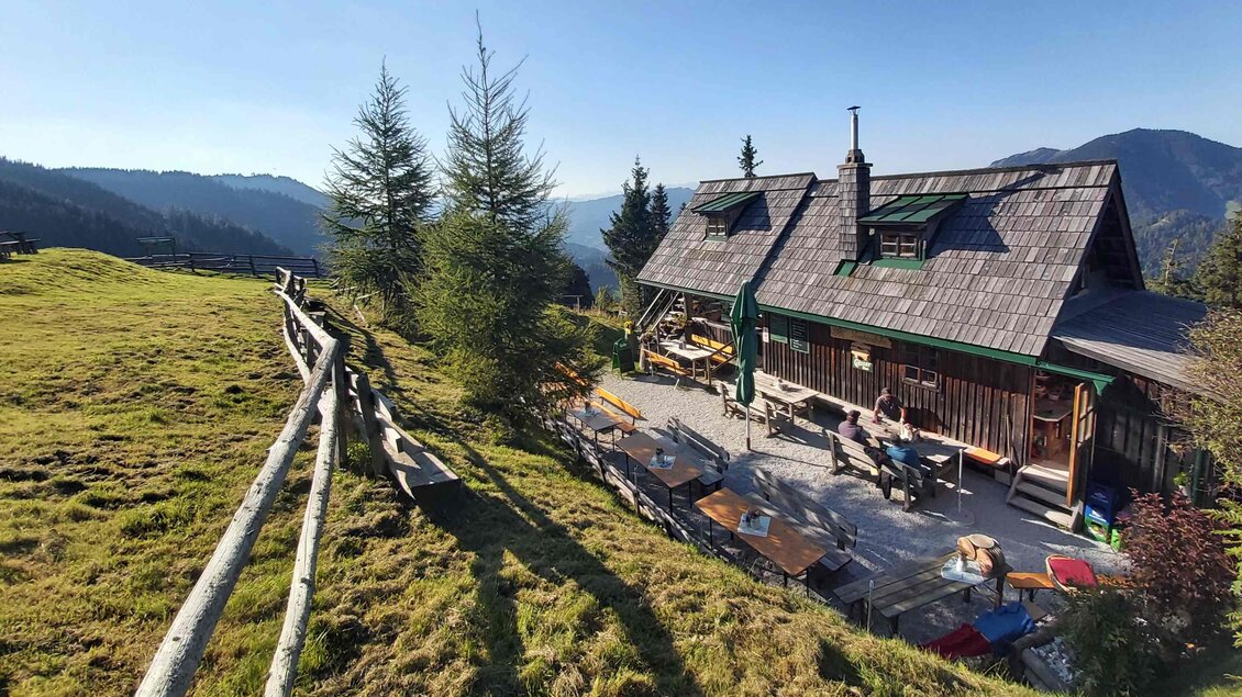 Eine malerische Berghütte umgeben von Wiesen und Bäumen. Im Hintergrund sind sanfte Hügel und blauer Himmel zu sehen. | © Oststeiermark Tourismus