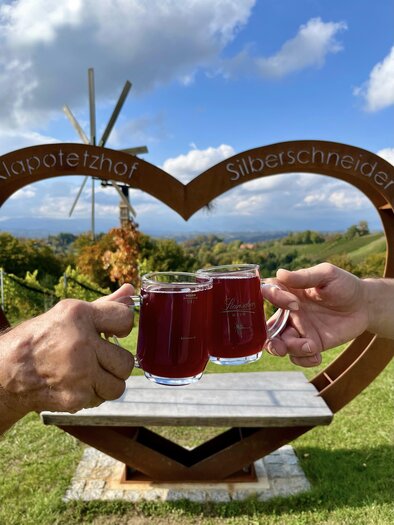 Two hands clink with mugs filled with red drink. In the background, there is a heart-shaped structure with a beautiful landscape and a wind turbine visible. | © Klapotetzhof Silberschneider