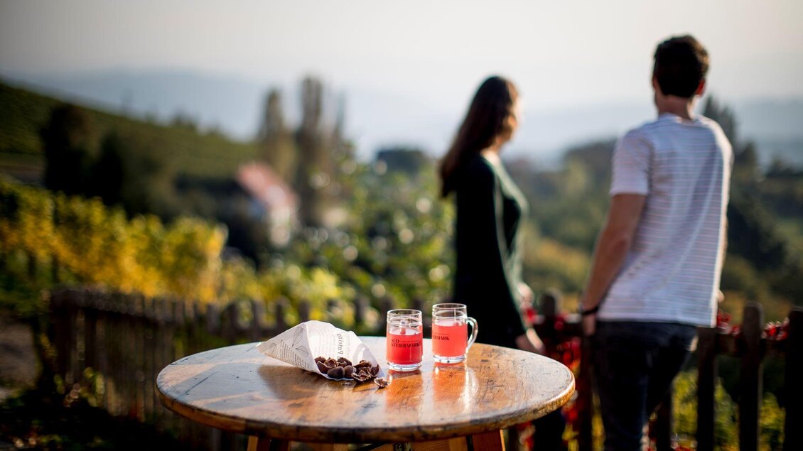 Ein Tisch mit zwei Gläsern Getränk und einer Dekoration aus Früchten steht im Vordergrund. Im Hintergrund sind zwei Personen zu sehen, die die schöne Landschaft genießen. | © TV Südsteiermark - Tom Lamm