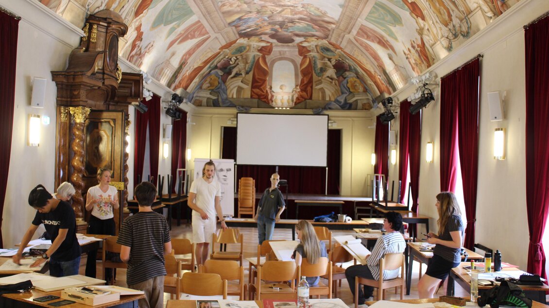 Ein heller Seminarraum mit kunstvollen Deckenfresken und roten Vorhängen. Gruppen von Menschen arbeiten an Tischen und scheinen an einem kreativen Projekt zu sein. | © Christian Strassegger