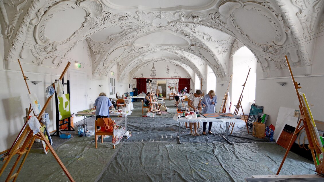 Ein großes, helles Atelier mit künstlerischen Arbeiten und mehreren Malstaffeleien. Menschen sind beschäftigt, verschiedene Kunstwerke zu schaffen. | © Christian Strassegger