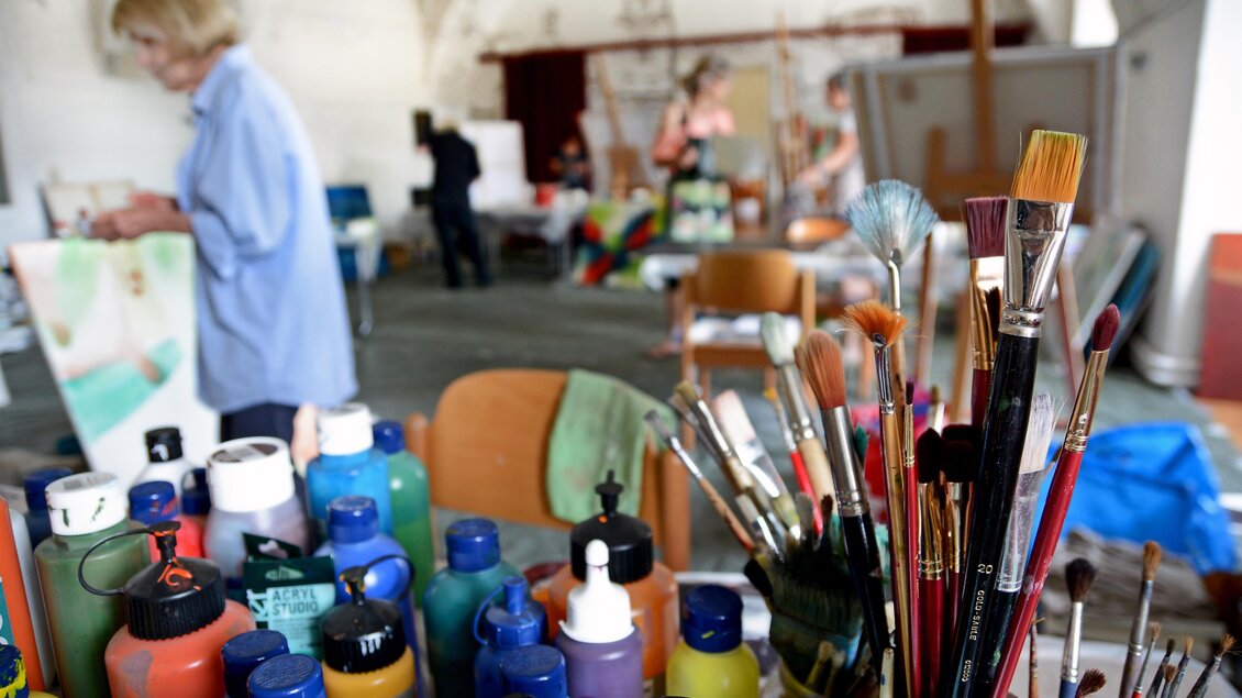 Eine kreative Atelierumgebung mit Pinsel und Farben im Vordergrund. Im Hintergrund arbeiten Personen an ihren Kunstwerken. | © Christian Strassegger