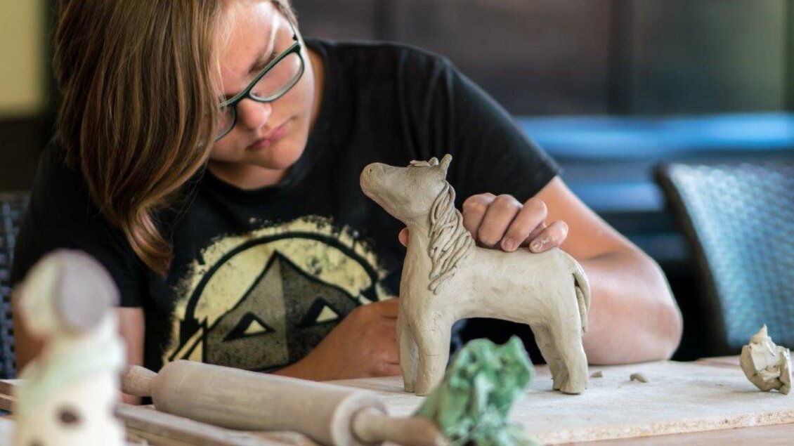 Ein junger Mensch formt eine Pferdefigur aus Ton. Die Umgebung zeigt eine kreative Werkbank mit Werkzeugen und Materialien. | © Christian Strassegger
