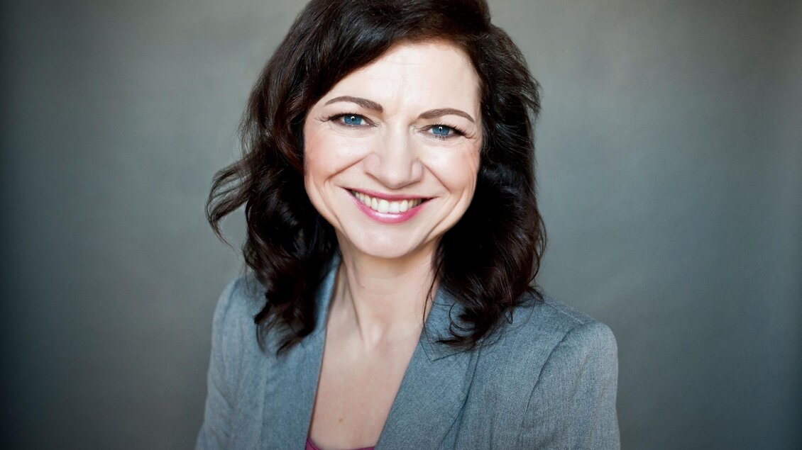 Eine Frau mit lockigem, dunklem Haar lächelt freundlich in die Kamera. Sie trägt einen grauen Blazer und hat einen hellen Hintergrund. | © Miriam Raneburger