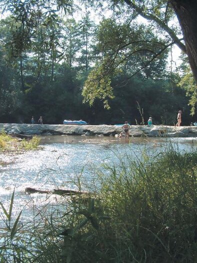 Ein ruhiger Fluss mit schimmerndem Wasser und umgebenden Bäumen. Im Hintergrund sind Menschen, die im Wasser entspannen und spielen. | © Steinerne Wehr