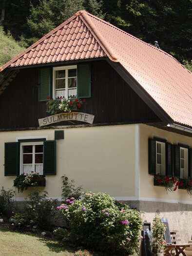 Ein charmantes Haus mit einem roten Dach und grünen Fensterläden. Es liegt in einer malerischen Landschaft, umgeben von Bäumen und Blumen. | © nadinegeuterphotography